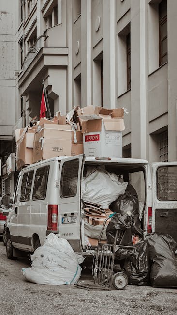 A white panel van parked on a city street adjacent to a tall, multi-storey building with a light-colored facade and architectural details. The van's rear doors are open, revealing an interior filled with large black garbage bags, cardboard boxes, and packing materials related to home relocation and furniture transport. Several cardboard boxes, some sealed with packing tape, are stacked on top of the van, with labels visible on some. A trolley is positioned near the open back of the van, indicating the loading process for moving debris and unwanted items following a house move. The scene is set during daylight with natural lighting, capturing a typical bulky waste removal operation coordinated by [COMPANY_NAME], which specializes in removals and relocation services in Marylebone. The surroundings include pavement and part of the building’s exterior, illustrating the typical logistical steps involved in packing, loading, and transporting household items during a house move. This image exemplifies the final stage of packing and moving, highlighting aspects of bulky waste removal after a home relocation.