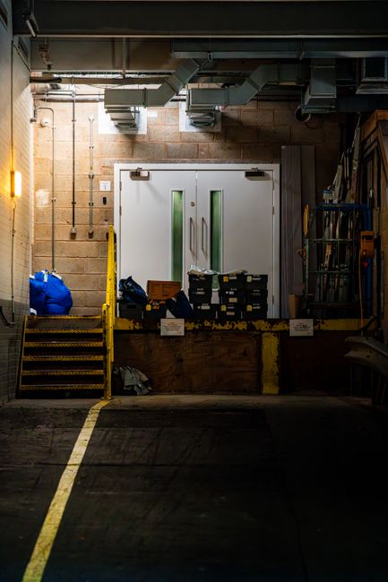 Inside a loading bay area of a building, a white double door with small rectangular windows is visible, set against a concrete brick wall. To the left of the door, a yellow safety barrier and a metal 