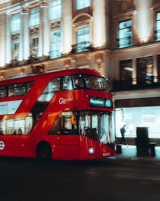 A red double-decker London bus traveling along a city street at night, with illuminated historic building facades featuring large windows, decorative columns, and ornate architectural details in the b