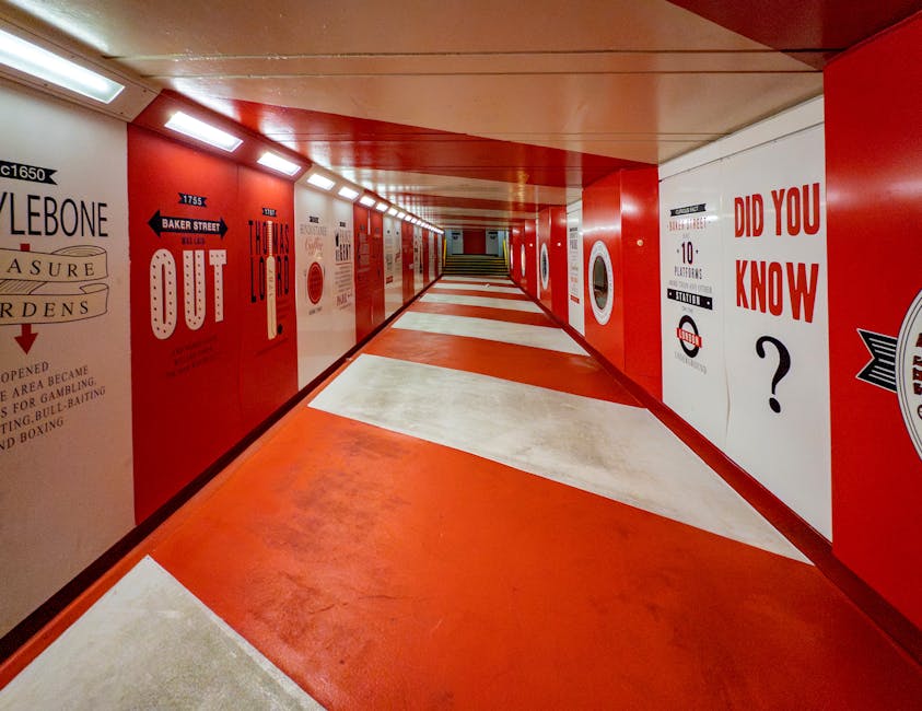 A long underground pedestrian tunnel with red and white walls featuring various signs and advertisements, including a large red sign that reads 'OUT' with an arrow pointing to the left, and other smaller text advertisements for local venues. The floor has a striped pattern with red and white sections, and the ceiling is fitted with rectangular fluorescent lights. This environment is part of the internal route used for moving or transporting furniture and boxes during a home relocation, with the tunnel providing access between different parts of a building or to an exit point. The scene is well-lit, and the space appears clean and unobstructed, supporting smooth loading, unloading, or transport activities consistent with professional removal services such as those offered by Removals Marylebone.