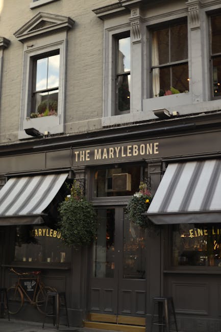 Exterior view of a two-story building on Marylebone High Street with a black sign reading 'THE MARYLEBONE' above the entrance, which is flanked by striped black and white awnings. The facade features large windows with white frames on the upper floor and potted plants hanging beside the door. The street scene includes a small outdoor seating area with a wooden chair and table to the left of the entrance. This setting depicts a commercial property, possibly a cafe or pub, with a focus on the building's traditional architectural details. The image is related to house removals and moving services, highlighting the street environment where residential and commercial relocations may occur, and is hosted on https://removalsmarylebone.org.uk under the 'Marylebone High Street Moves: Best Times & Parking W1U, Marylebone' page.