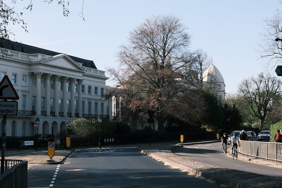 A wide view of Montagu Square in Marylebone during daytime, featuring a large white neoclassical building with columns on the left, and a prominent domed building in the distance. The scene includes a tree with sparse foliage in the center, indicating early spring or late winter, and a paved road with a gentle curve leading towards the background. On the right, several cyclists and pedestrians are moving along the pavement past a metal railing. The area is illuminated by natural light, with shadows cast by the trees and structures, and traffic cones and signage indicating active roadwork or traffic control. The scene captures an urban environment suitable for house removals or furniture transport, with typical street features such as parked cars, streetlamps, and directional signs, suggesting the logistical considerations involved in home relocation and packing within the Marylebone district, supported by Removals Marylebone.