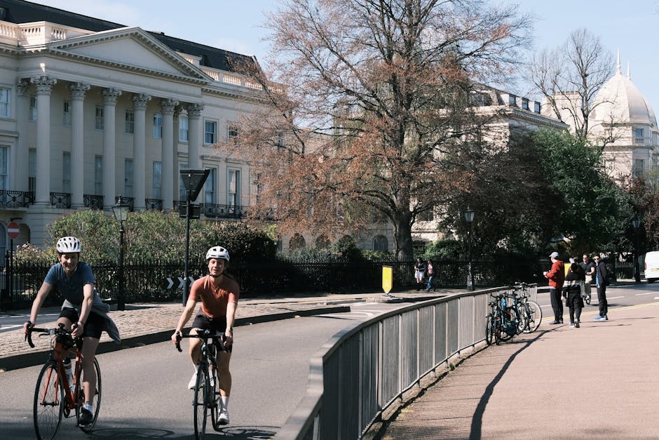 A wide view of Montagu Square in Marylebone during daytime, featuring a large white neoclassical building with columns on the left, and a prominent domed building in the distance. The scene includes a tree with sparse foliage in the center, indicating early spring or late winter, and a paved road with a gentle curve leading towards the background. On the right, several cyclists and pedestrians are moving along the pavement past a metal railing. The area is illuminated by natural light, with shadows cast by the trees and structures, and traffic cones and signage indicating active roadwork or traffic control. The scene captures an urban environment suitable for house removals or furniture transport, with typical street features such as parked cars, streetlamps, and directional signs, suggesting the logistical considerations involved in home relocation and packing within the Marylebone district, supported by Removals Marylebone.