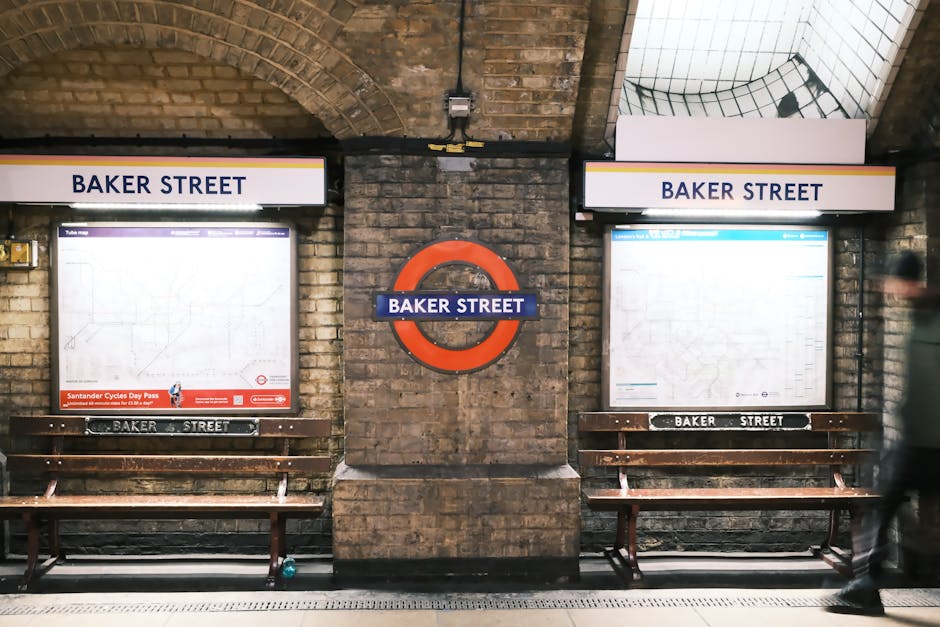 Inside Baker Street underground station, two illuminated station maps are mounted on a brick wall with a central roundel sign displaying 'BAKER STREET'. Each map is framed and positioned above wooden benches with 'BAKER STREET' printed on a metal strip beneath each. The left map includes advertisements at the bottom, and both maps display local transit routes. To the right, a blurred figure is walking past, indicating transit activity. Overhead, the arched brick ceiling is visible along with some wiring and grid panels, emphasizing the station's historic architecture. The setting is well-lit with natural and artificial lighting, capturing the typical environment of home relocation and furniture transport logistics associated with moving services such as those offered by Removals Marylebone. This scene exemplifies the typical loading and loading process at a London tube station, relevant to urban house removals and packing operations.