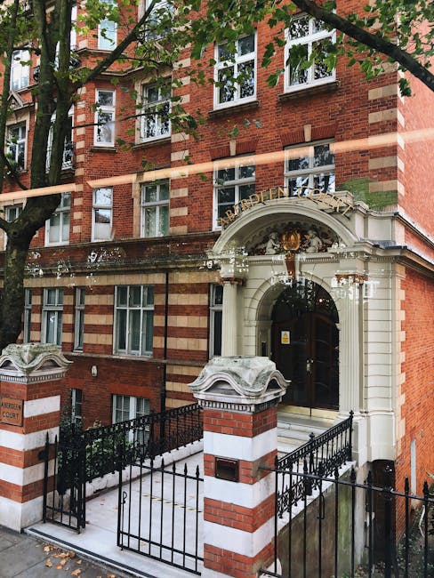 The entrance to a multi-story red brick residential building in Marylebone, featuring white stone accents around the doorway and a decorative curved arch with a gold crest above the door. There are two brick pillars with white stone caps supporting a black iron gate, which encloses a small front courtyard. The building has several white-framed windows with mullions aligned vertically and horizontally. A large tree with green leaves partially shades the facade, and sunlight filters through the branches, creating dappled light on the building. The scene reflects a typical urban setting suitable for home relocation or furniture transport services, with the building's entrance prepared for a residential move by [COMPANY_NAME], highlighting the accessibility and logistical considerations involved in house removals in Marylebone.