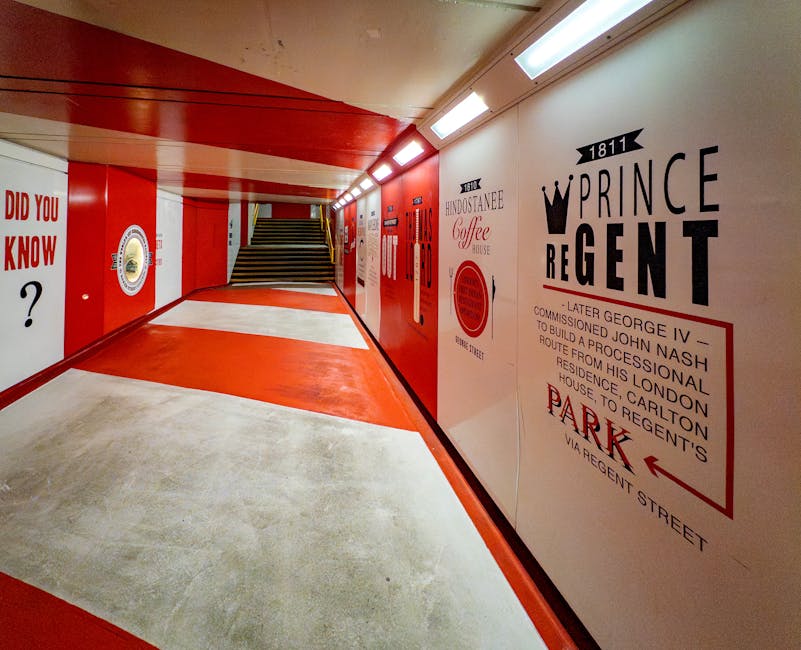 Inside a brightly lit underground pedestrian tunnel with red and white painted walls, an individual is seen lifting or carrying a cardboard box wrapped in plastic film, likely for home relocation or furniture transport. The floor features a combination of concrete and a red painted pathway, guiding towards a staircase at the end of the corridor. The walls are decorated with printed advertisements and historical information in black and red typography, including references to The Prince Regent and the East End of London. Fluorescent ceiling lights illuminate the space, creating a well-lit environment for moving activities. This scene exemplifies the loading process during house removals, with boxes and possibly small furniture items prepared for transport by [COMPANY_NAME], situated within a building accessed via stairs. The environment suggests a professional moving operation focused on meticulous handling of packing materials and efficient navigation through indoor spaces.
