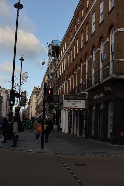 The entrance to a multi-story red brick residential building in Marylebone, featuring white stone accents around the doorway and a decorative curved arch with a gold crest above the door. There are two brick pillars with white stone caps supporting a black iron gate, which encloses a small front courtyard. The building has several white-framed windows with mullions aligned vertically and horizontally. A large tree with green leaves partially shades the facade, and sunlight filters through the branches, creating dappled light on the building. The scene reflects a typical urban setting suitable for home relocation or furniture transport services, with the building's entrance prepared for a residential move by [COMPANY_NAME], highlighting the accessibility and logistical considerations involved in house removals in Marylebone.