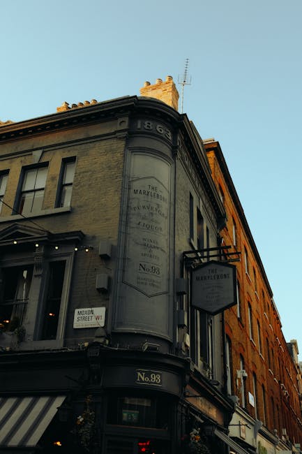 A street scene in Marylebone with a multi-storey brick building featuring arched windows and balconies on the right side, partially covered by scaffolding for maintenance or renovations. The ground fl